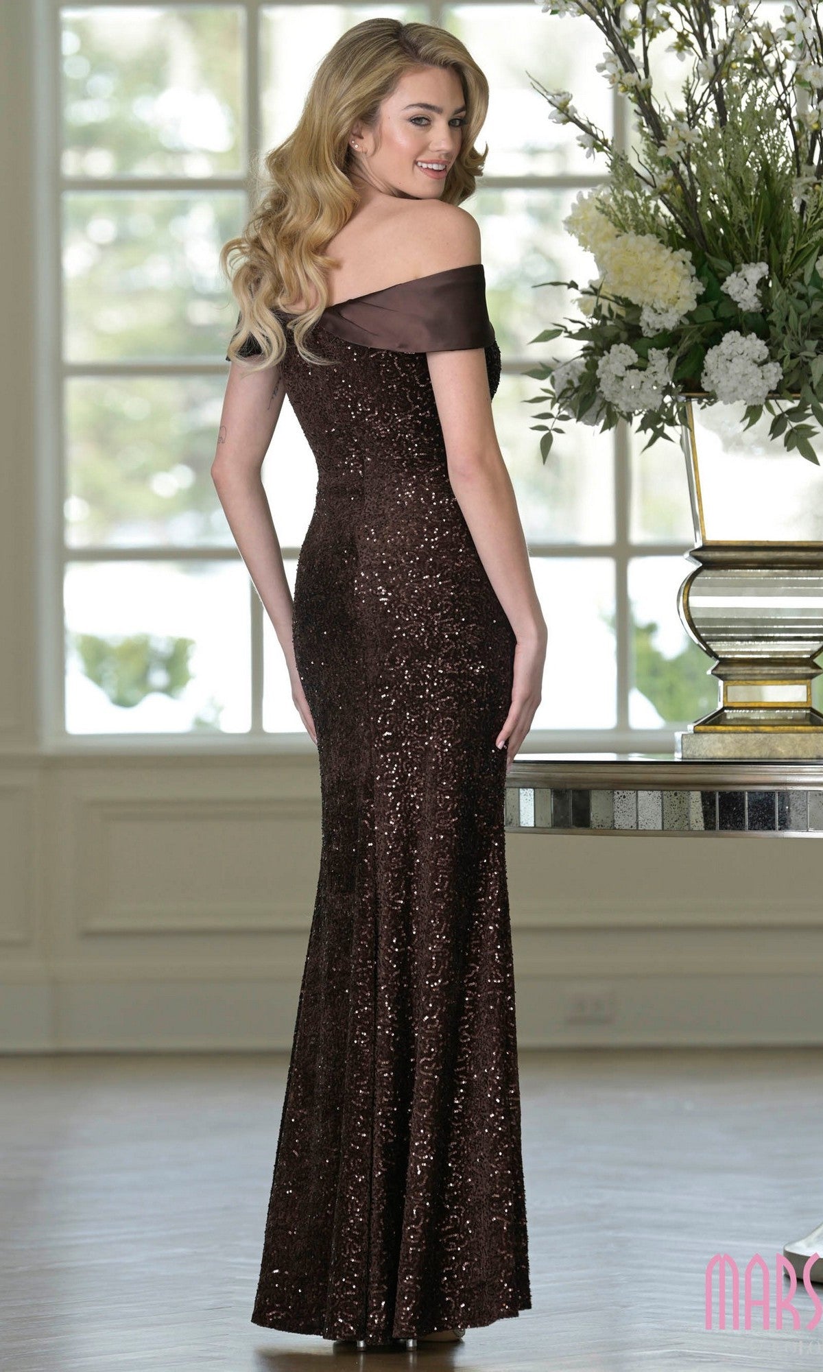 A woman with long wavy hair stands indoors near a large floral arrangement, wearing the Marsoni M349 Long Formal Dress, a floor-length, off-the-shoulder gown adorned with dark brown sequins.